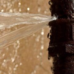 A rusty metal pipe with a significant leak, spraying water forcefully from its side against a blurred background.