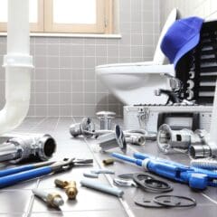 A bathroom with gray tiles, featuring plumbing tools and parts scattered on the floor. Tools include wrenches and pipes. A tool case is open with a blue cap resting on it. A toilet is in the background near a window.
