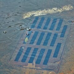 Rainwater with ripples surrounds a partially submerged street drain in an asphalt surface. The metal grating is visible under the water, and small circular ripples indicate falling raindrops. A leaf is caught in the water near the drain.
