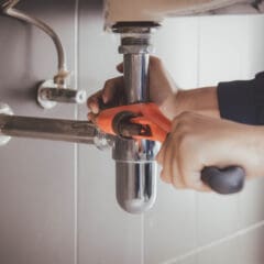 Hands using a red pipe wrench to tighten or loosen a shiny metal pipe under a sink. The background features gray wall tiles and additional plumbing components.