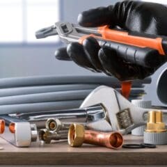 A person wearing a black glove holding an adjustable wrench, surrounded by various plumbing tools and materials, including copper pipes, fittings, and hoses, on a table. Coiled tubing is in the background, with a blurred window view.