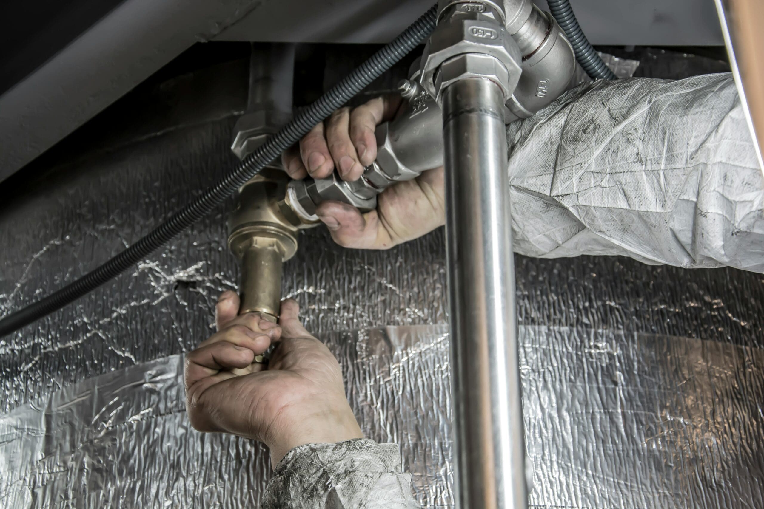 Person wearing protective clothing, using their hands to tighten or adjust a metal pipe under a sink or appliance. The setting appears industrial or maintenance-related, with reflective foil insulation in the background.