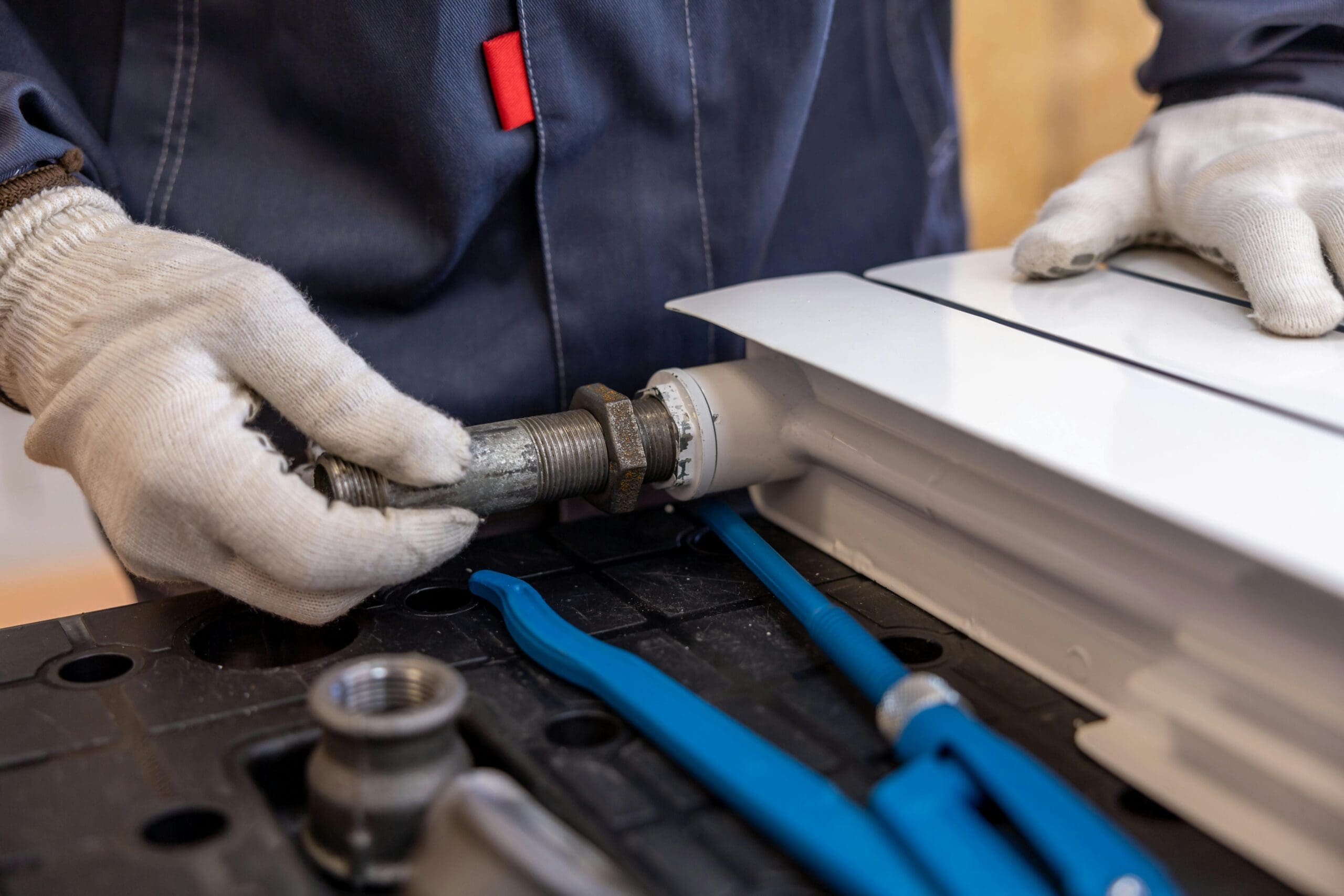 Person wearing gloves and a dark jacket connects piping fixtures with tools on a table, working on a white appliance.