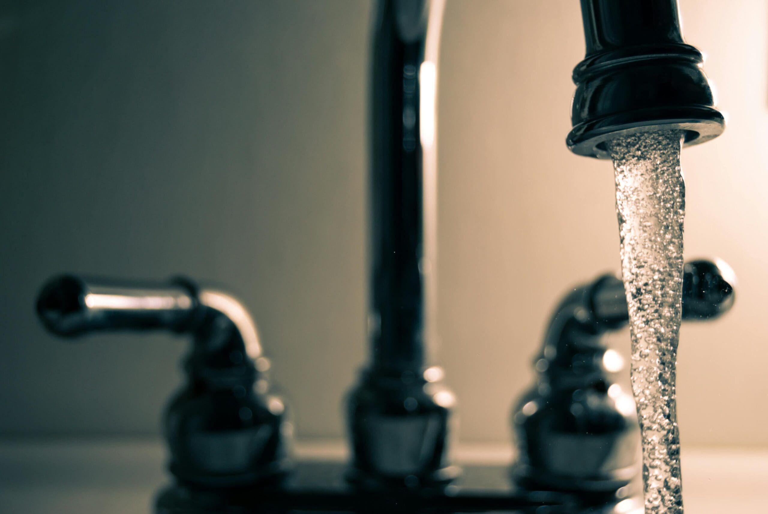 Close-up of a bathroom faucet with a sleek design as water flows steadily from the spout. The faucet features two handles for controlling temperature. The background is softly blurred, highlighting the polished metal surface against a warm light.