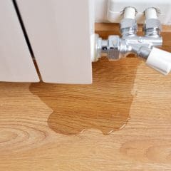 A small puddle of water is forming on a wooden floor beneath a radiator valve, indicating a leak. The wood grain is visible, and the radiator is white with metal and plastic connections.