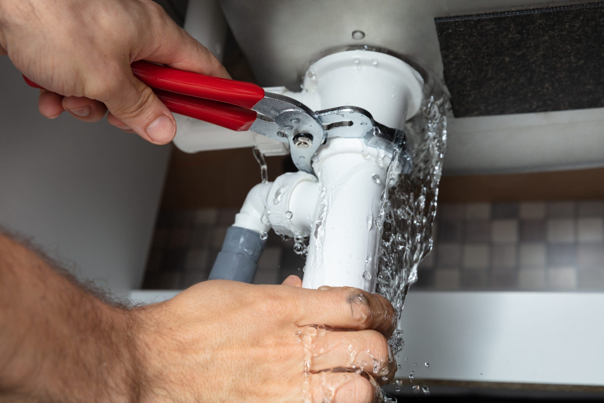 Person using red-handled pliers to fix a leaking white pipe under a sink. Water is spraying from the pipe as the person attempts to tighten the connections. The background has a tiled pattern.