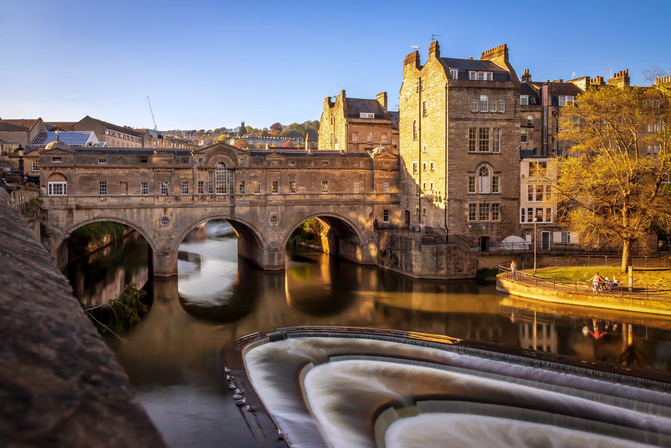 Historic stone bridge with arches spans over a river in an old town, bathed in warm sunlight. There are classic buildings on the riverbank and calm water flow, with a few people sitting by the riverside. Blue sky forms the backdrop.