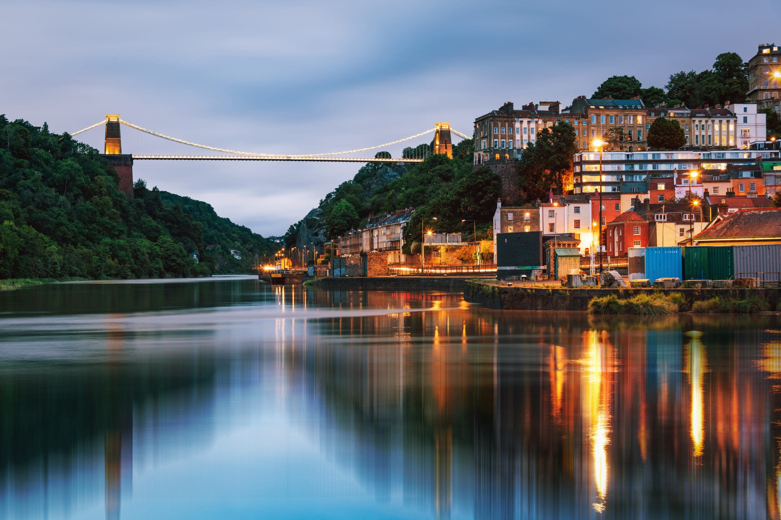 View of Clifton Suspension Bridge illuminated at dusk, spanning over a calm river. Houses and buildings climb the hill on the right, reflecting in the water. Lush greenery surrounds the scene, with a cloudy sky above.