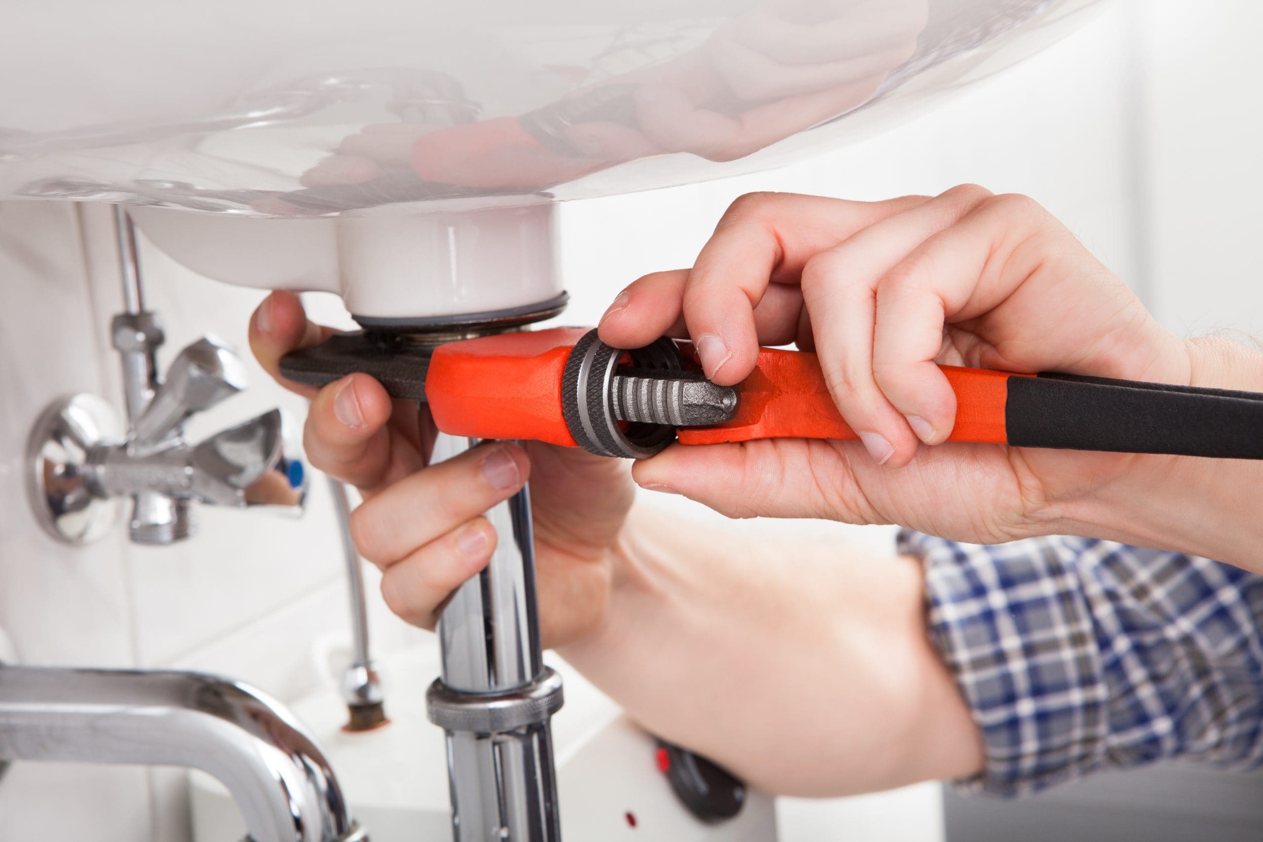 Close-up of a person using an orange wrench to tighten or loosen a pipe under a sink. The hands are focused on the task, and the sink's plumbing is visible in the background.