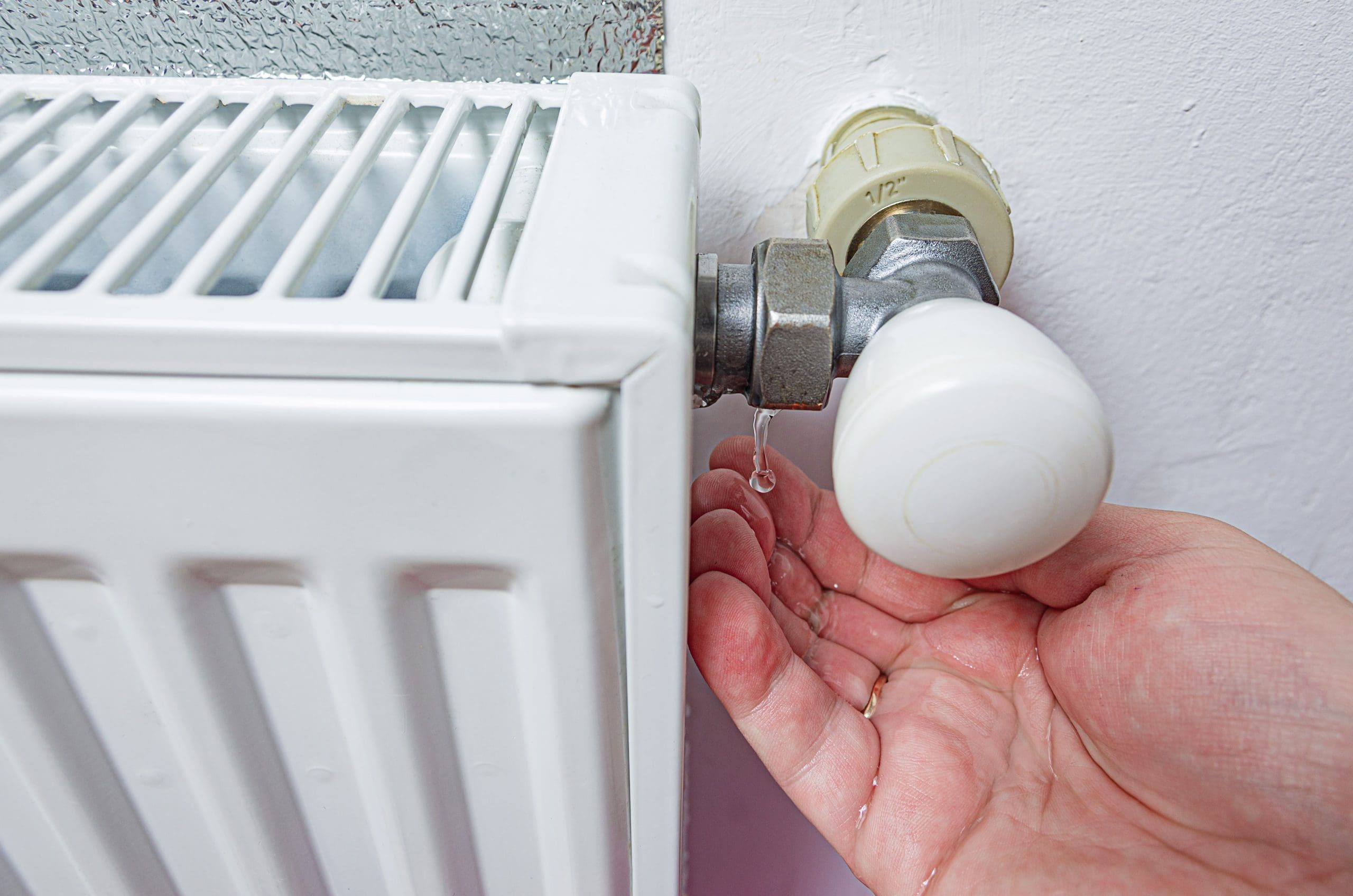 A hand checks for a water leak under a white radiator valve against a white wall. The valve is silver with a white cap, and water is dripping from it.