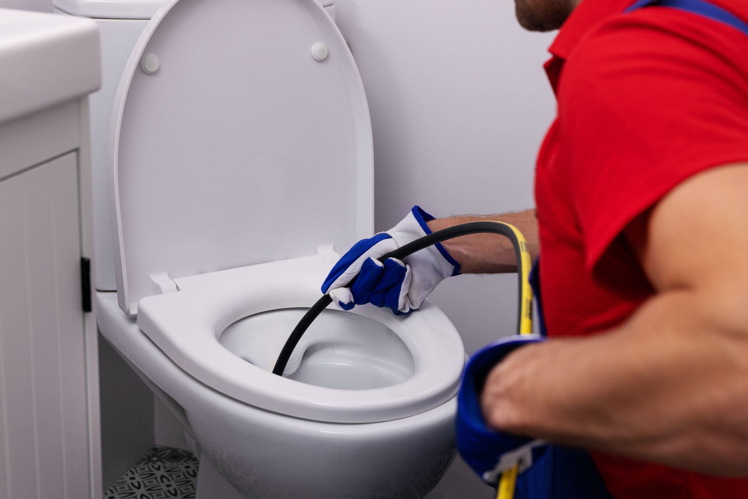A plumber in a red shirt and blue gloves is using a plumbing snake to unclog a white toilet in a bathroom. The partial view shows the plumber's torso and arms while focusing on the cleaning process.