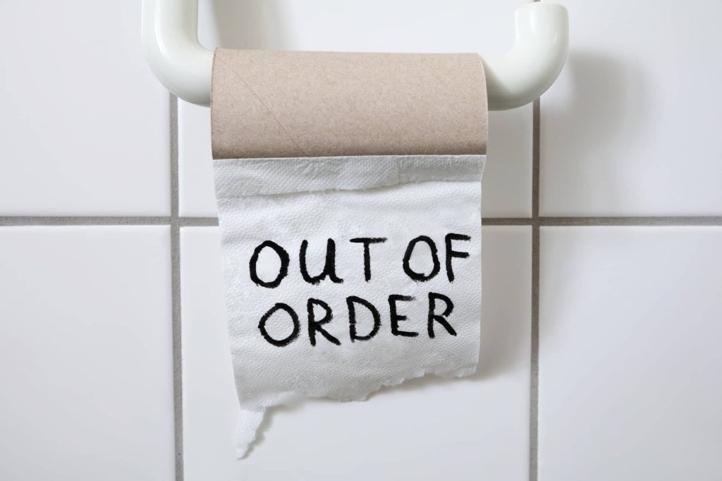 A toilet paper roll hung on a holder, with the last sheet hanging down and the words "OUT OF ORDER" written on it in black marker. The roll is empty, against a white tiled wall.