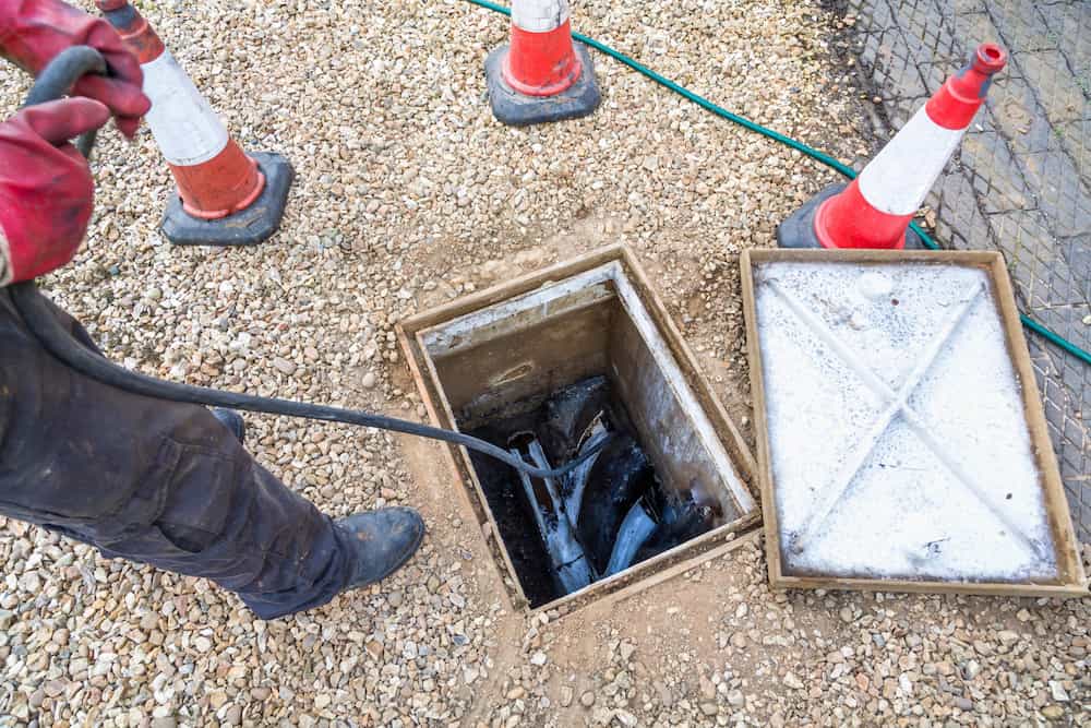 A person wearing red gloves is handling a cable near an open utility manhole. The manhole cover is removed and placed to the side. Traffic cones surround the area for safety.