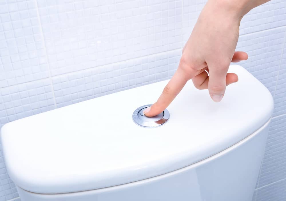 A hand pressing the flush button on top of a white toilet tank against a tiled bathroom wall.